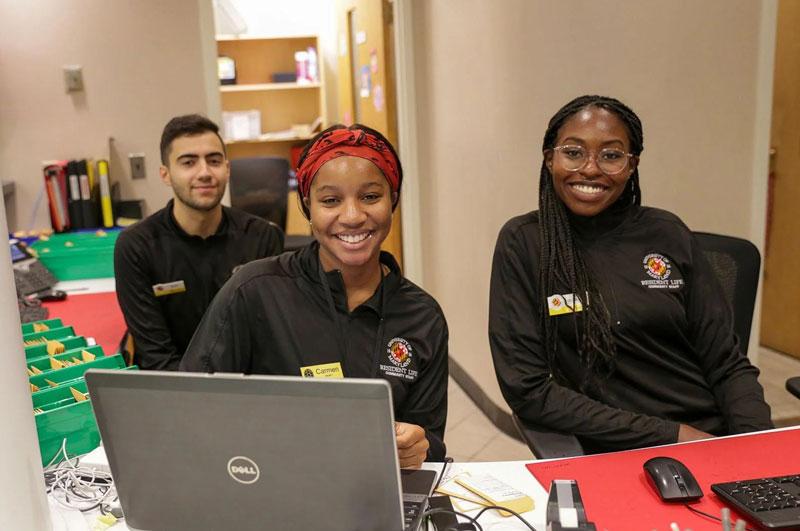 three community assistants behind a desk in Annapolis Hall smiling at camera